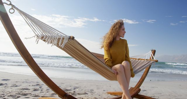 A young Caucasian woman relaxes in a hammock on a sunny beach, with copy space. Her serene expression and the tranquil seaside setting evoke a sense of peace and leisure.