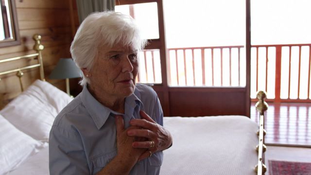 Senior woman clutching her chest in discomfort, possibly indicating a health issue. Scene is set in a well-lit bedroom with a wooden interior. Suitable for use in health-related articles, senior care advisories, emergency awareness campaigns, or medical blogs focusing on elderly health issues.