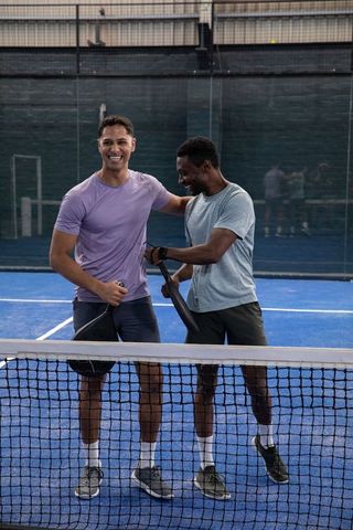 Smiling male friends celebrating on blue padel court at net showing wristwatch and camaraderie