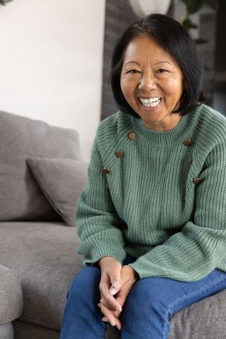 Smiling Senior Asian Woman with Green Sweater on Grey Sofa
