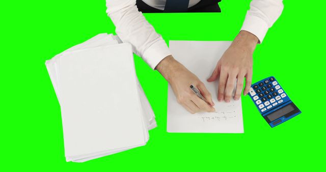 A Caucasian middle-aged businessman is writing on a sheet of paper, with copy space on the green background. The presence of a calculator and stack of papers suggests financial work or accounting tasks.