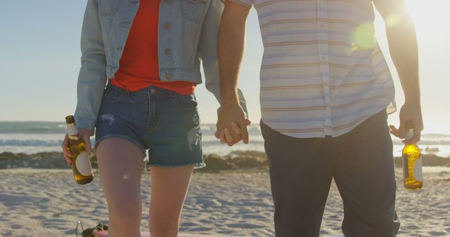 Mid section of couple holding hands with beer bottle walking on beach - Download Free Stock Photos Pikwizard.com