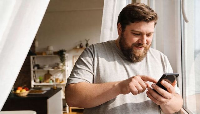 Bearded man texting on smartphone by sunlit window in cozy kitchen
