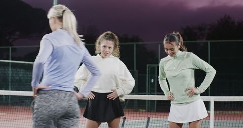Female tennis players stretching and chatting by net at twilight on outdoor court