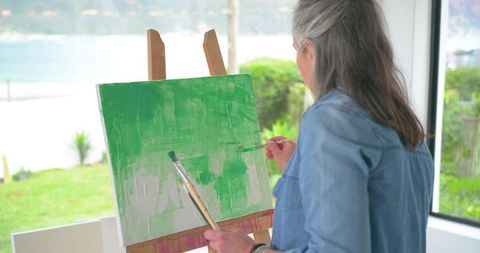 Senior woman painting green abstract on canvas at bright seaside studio with brushes and easel