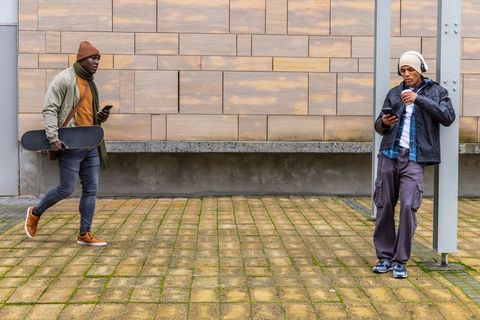 Urban youth walking with skateboard and coffee using phones in overcast courtyard plaza