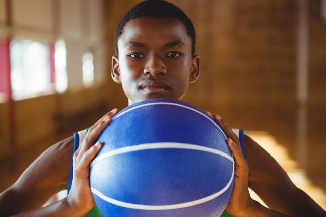 Young man holding a blue basketball with a serious expression in an indoor court. Ideal for use in sports-related content, youth athletic programs, fitness promotions, and motivational materials.