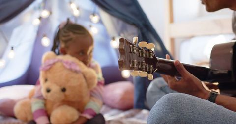 Dad playing acoustic guitar while child hugging teddy in cozy teepee fort
