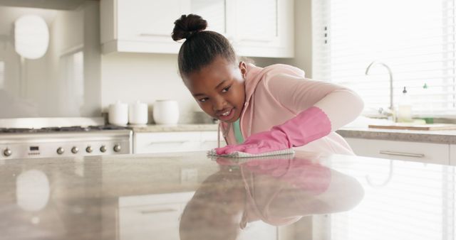Young girl wearing pink gloves cleaning kitchen counter. Useful for content related to household chores, hygiene education, cleaning products, child responsibilities.