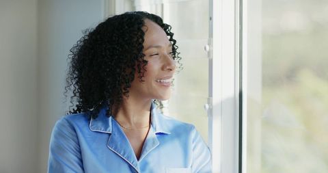 Woman in blue pajamas standing by window enjoying morning sunlight and calm reflection