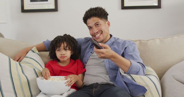 Father and young daughter sitting on a beige couch in the living room. Father is using a remote control, both relaxed and casual, bonding over a TV show. The girl is wearing a red shirt and is holding a bowl of popcorn. The scene is a perfect representation for advertisements and content related to family time, parenting, leisure, or home decor.
