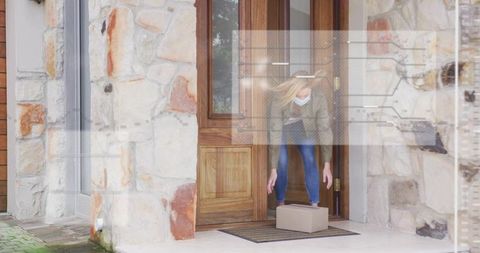 Masked woman picking up package from doorstep of stone facade home during delivery