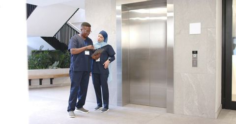 Diverse clinicians consulting on tablet in modern hospital lobby by elevator and stairs