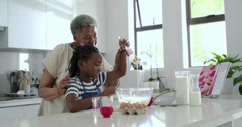 African american grandmother guiding granddaughter mixing batter in sunlit modern kitchen