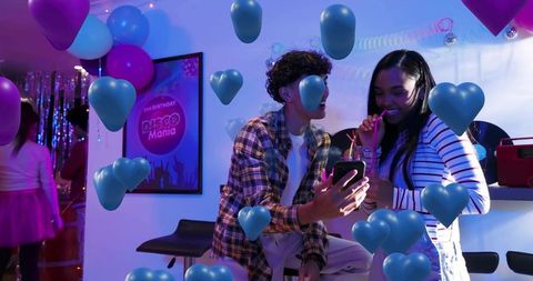 Sharing smartphone at neon disco lounge with floating heart balloons and colorful lighting