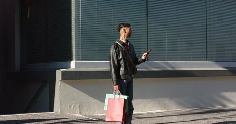 Young man checking phone and carrying shopping bags on sidewalk in urban sunlight