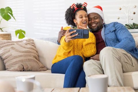 African american couple taking cozy christmas selfie on couch wearing santa hats, smiling