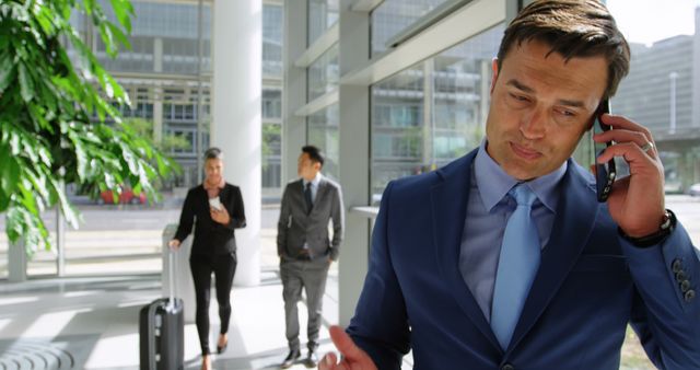 A Caucasian middle-aged businessman in a blue suit is on a phone call, with copy space. In the background, a diverse group of professionals is walking through a modern office building's lobby.