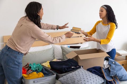 Mother and teen daughter packing and sorting clothes while exchanging books in bedroom
