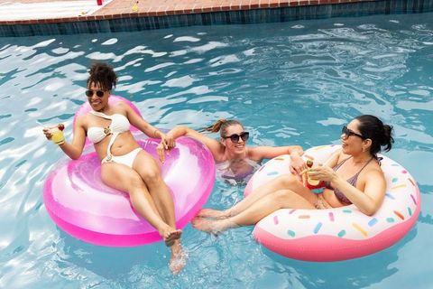 Diverse women relaxing on inflatable donut floats in backyard pool sipping cocktails