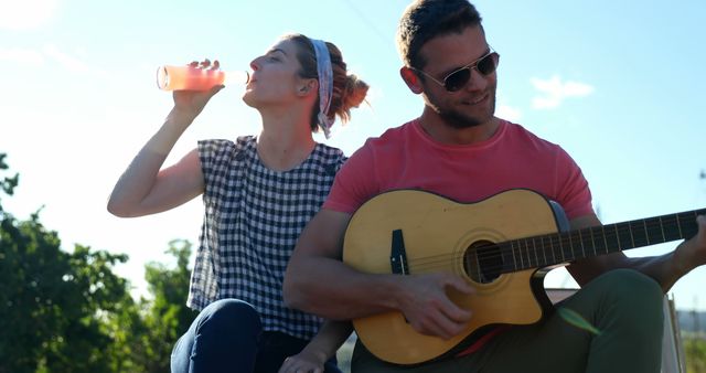 Caucasian couple enjoys music outdoors. He plays the guitar while she hydrates on a sunny day.