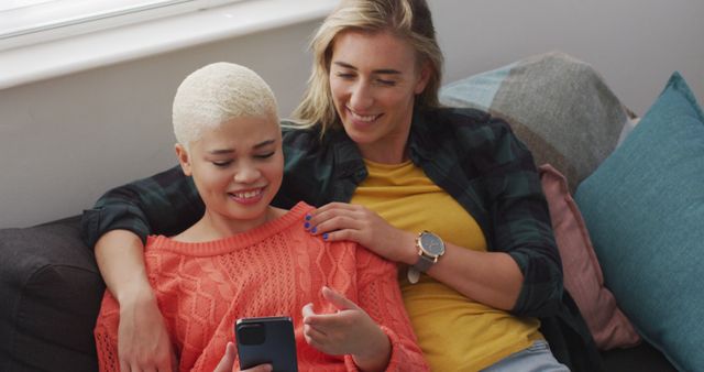 Two women are relaxing on a sofa, smiling while looking at a smartphone. This image is suited for contexts such as online articles about modern relationships, lifestyle blogs, social media promotions, or advertisements focusing on home technology, leisure, and diversity.