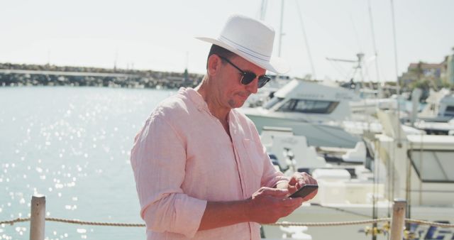 Happy caucasian man in hat and sunglasses using smartphone on jetty by boats on a sunny day. Leisure, free time, communication, travel and vacations.