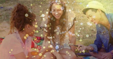 Three friends laughing on picnic blanket sharing smartphone during golden hour