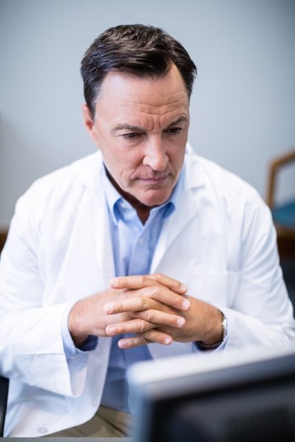 Male doctor in white coat concentrating on computer screen in clinic. Ideal for use in healthcare, medical technology, telemedicine, and patient care contexts. Can be used in articles, advertisements, and educational materials related to medical professionals and healthcare settings.