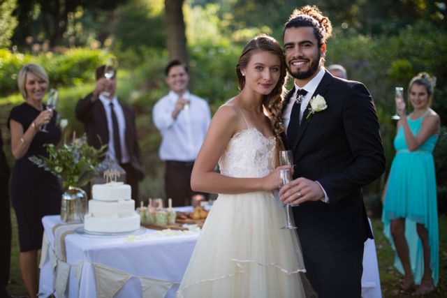 Newly married couple celebrating their wedding with friends in a park. The bride and groom are holding glasses of champagne, standing in front of a decorated table with a wedding cake. Friends are toasting in the background. Ideal for use in wedding planning materials, celebration announcements, and romantic event promotions.