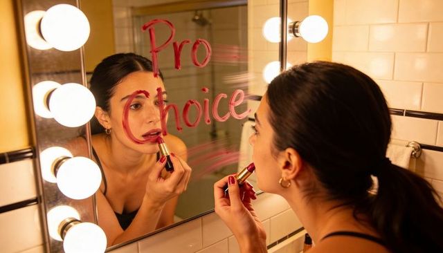 Young woman writing pro choice with lipstick on vanity mirror backlit by hollywood lights