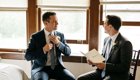 Groom and groomsman adjusting ties while reading vow program in sunlit room