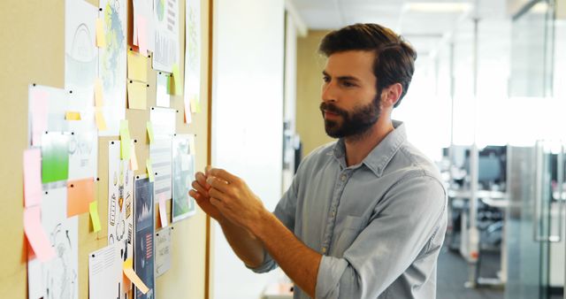 The image shows a man in an office sticking notes and papers on a bulletin board, indicating he is brainstorming and planning a project. This photo can be useful for articles or website content related to workplace organization, project management, creative processes, and team collaboration.