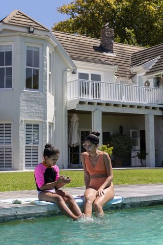 African mother and daughter sitting poolside wearing swimwear sharing seashell and summertime bond