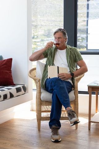 Mature man sipping tea and journaling in sunlit wicker chair near window