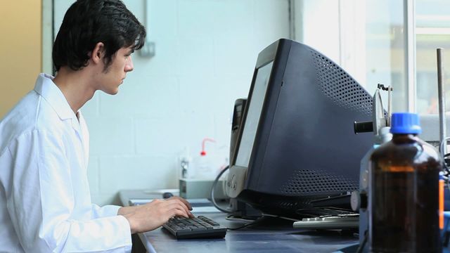 Young male scientist sits at a desk in a laboratory, intensely working on a desktop computer. Dressed in a white lab coat, he is engaged in scientific research or data analysis. The scene portrays an environment of scientific inquiry and technological advancement, making it ideal for illustrating themes of scientific research, laboratory work, technology in science, or professional dedication in educational or promotional materials.