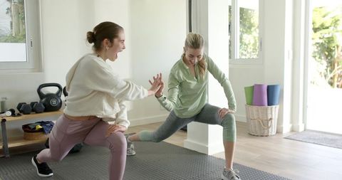 Female friends lunging and high-fiving during home workout in bright living room