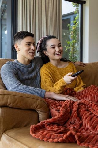 Diverse couple relaxing on leather sofa watching tv laughing under cozy blanket