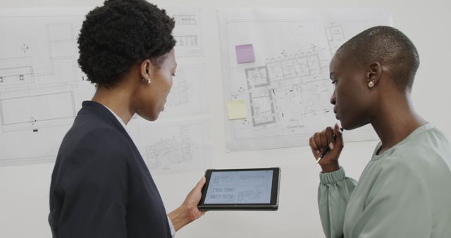 Two architects collaboratively analyzing building plans displayed on a tablet. The background shows large blueprints pinned to a wall. This image can be used for concepts related to engineering, architecture, teamwork, and modern technology in design logistics.