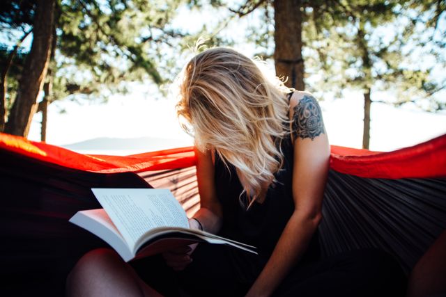 Woman with blonde hair and tattoo relaxing in hammock and reading book under trees. Ideal for illustrating concepts related to relaxation, outdoor activities, leisure, reading, and peaceful moments. Useful for blogs, articles, advertisements promoting self-care, mental well-being, and outdoor lifestyle.