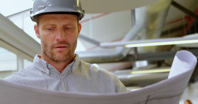 Engineer wearing hard hat carefully examining blueprint in modern office, suitable for illustrating concepts of engineering, construction projects, project planning, and professional work environments.