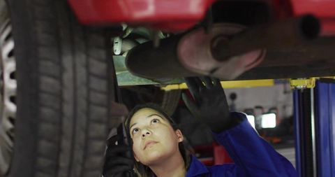 Female mechanic inspecting undercarriage of red car on lift wearing blue coveralls