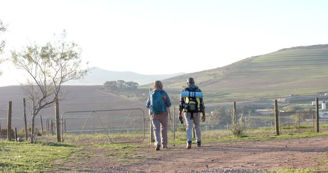 This image shows two hikers walking in the rural countryside, carrying backpacks and other hiking gear. The area is open and natural, with hills in the background and a dirt path underfoot. Ideal for use in travel blogs, adventure magazines, eco-tourism campaigns, or outdoor recreational advertisements.