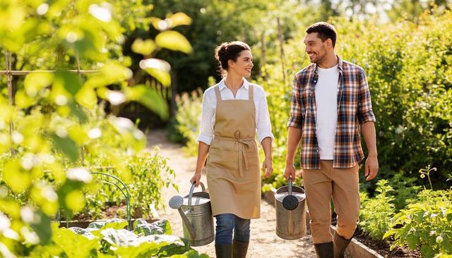 Couple walking through sunlit vegetable garden carrying watering cans, smiling