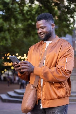 African man standing in evening city checking smartphone wearing orange bomber jacket