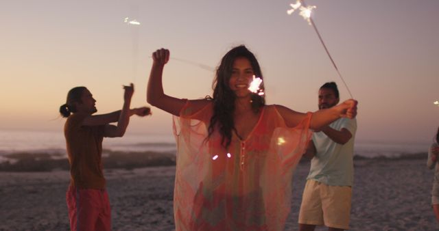 Happy caucasian woman having fun with diverse friends dancing on the beach with sparklers at sunset. Summer, free time, friendship, party, celebration and vacations.