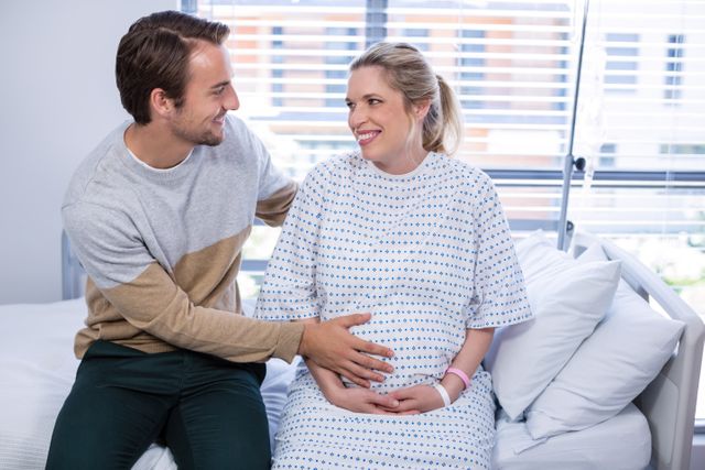 Man comforting pregnant woman in hospital ward. Both are smiling and looking at each other, showing support and love. Ideal for use in healthcare, maternity, and family-related content, illustrating themes of support, love, and prenatal care.