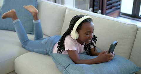African american preteen girl lying on sofa listening with headphones and using smartphone