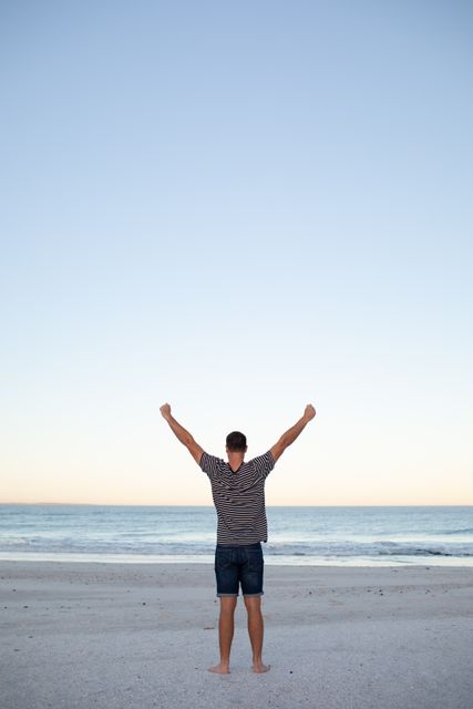 Man standing on sandy beach with arms raised, facing the ocean at sunrise. Ideal for concepts of freedom, success, and relaxation. Suitable for travel advertisements, motivational posters, and wellness blogs.