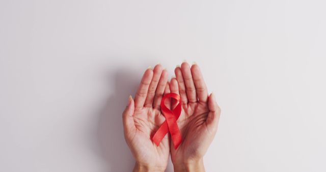 This image features hands gently holding a red ribbon, symbolizing AIDS awareness and support. It can be used for healthcare campaigns, educational materials, charity promotions, and events focused on HIV/AIDS awareness and prevention. Ideal for online articles, social media posts, medical newsletters, and promotional materials advocating for health and solidarity.